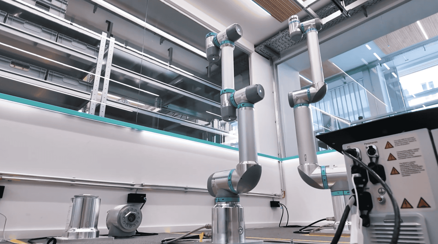 Two RobCo modular industrial robot arms standing in a bright, clean production facility with storage shelving and electrical control units.