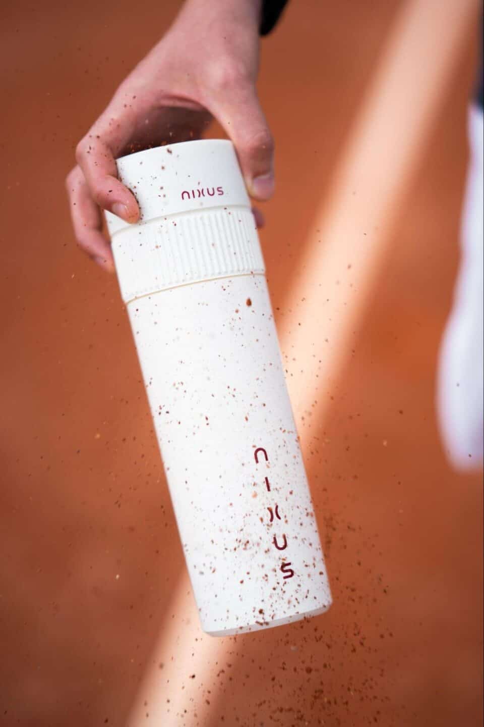 Action shot of a person holding the white NIXUS canister on a tennis court as red clay dust sprays across the frame.